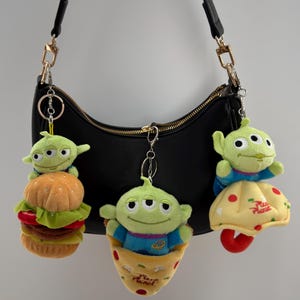 May include: A black shoulder bag with gold-colored hardware, showcasing three plush alien keychains. One alien sits atop a burger, another in a pizza slice, and the third on a Pizza Planet-themed item. The keychains are green, blue, and yellow.