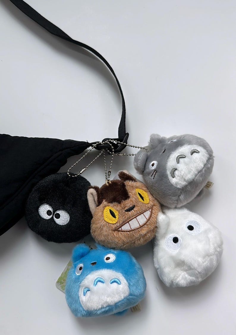 May include: A selection of plush keyrings featuring characters from a popular animated film. The keyrings include a black soot sprite, a brown cat-like creature, a grey Totoro, a white fluffy character, and a blue Totoro. They are attached to a black bag.