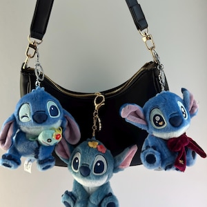 May include: Three blue plush Stitch keychains hanging from a black purse. Each keychain features a different Stitch design, with varying accessories like a small plush, flowers, and a red scarf. The purse has gold-colored hardware.