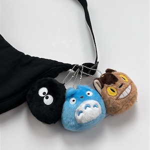 May include: Black crossbody bag with three plush charms. The charms include a black puff with white eyes, a blue character with a white belly, and a brown character with a yellow smile. The bag has a black strap.
