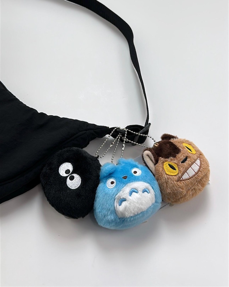 May include: Black crossbody bag with three plush charms. The charms include a black puff with white eyes, a blue character with a white belly, and a brown character with a yellow smile. The bag has a black strap.