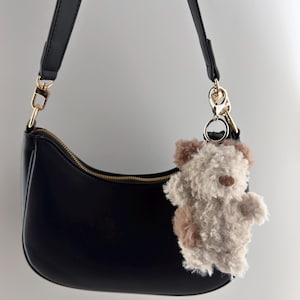 May include: A black, crescent-shaped handbag with a gold zipper and strap. A fluffy, teddy bear keychain in shades of brown and cream hangs from the strap. The bag is made of smooth material and has a simple, elegant design.