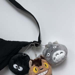 May include: A black crossbody bag with three plush character keyrings. The keyrings include a black soot sprite with white eyes, a brown cat-like creature with a wide grin, and a grey Totoro with white belly.