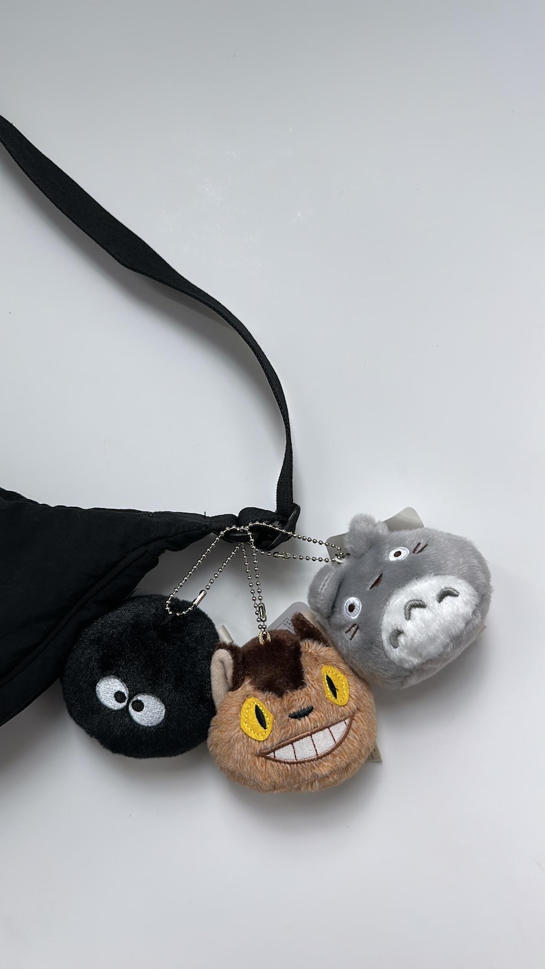 May include: A black crossbody bag with three plush character keyrings. The keyrings include a black soot sprite with white eyes, a brown cat-like creature with a wide grin, and a grey Totoro with white belly.