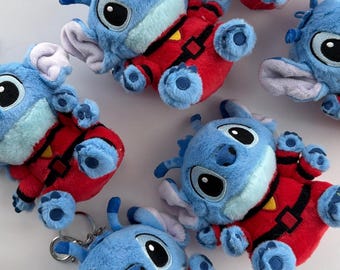 May include: A collection of blue plush toys featuring large ears, black eyes, and a light blue muzzle. Each plush character wears a red outfit with a black belt and a small yellow triangle detail on the chest. One plush includes a silver metal keychain ring.