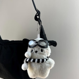 May include: A white plush toy with black aviator goggles and a black and white striped scarf. The toy is hanging from a black strap, and has black ears. The toy is a cute, whimsical accessory.