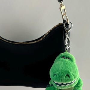 May include: A black shoulder bag with a gold-toned zipper closure and a black strap, featuring a plush green dinosaur keychain attached to the silver-toned metal hardware ring. The dinosaur has a wide, smiling mouth with visible white teeth.