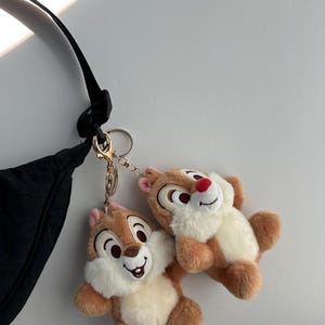May include: Two plush Chip and Dale keychains. The keychains feature the cartoon chipmunks with brown fur, white bellies, and red noses. They are attached to a gold-colored clasp and chain, hanging near a black bag.