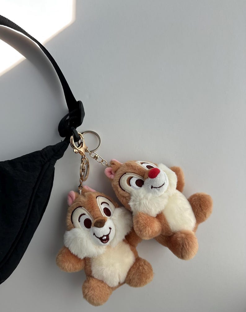 May include: Two plush Chip and Dale keychains. The keychains feature the cartoon chipmunks with brown fur, white bellies, and red noses. They are attached to a gold-colored clasp and chain, hanging near a black bag.