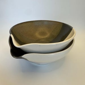 May include: Two ceramic bowls stacked on top of each other. The top bowl has a brown interior with a black and white exterior. The bottom bowl is white with a black and gray exterior. The bowls have a unique, asymmetrical shape.