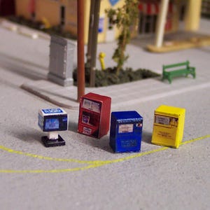 May include: Miniature models of a newsstand, a red vending machine, a blue vending machine, and a yellow vending machine. The models are on a grey surface with yellow painted lines, suggesting a street scene.