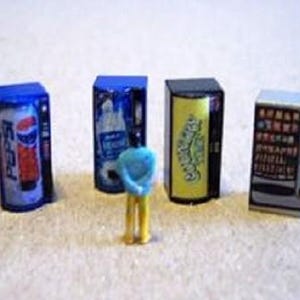 May include: Miniature vending machines, including a Pepsi machine, a water machine, a snack machine, and a candy machine. A small blue figure stands in front of the machines. The machines are rectangular with blue, black, and white color schemes.