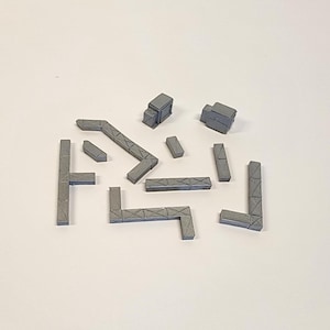 Puede incluir: Un conjunto de bloques de construcción geométricos grises en varias formas, incluyendo formas de L, T y prismas rectangulares. Los bloques están dispuestos sobre una superficie blanca, sugiriendo un tema de construcción o arquitectura.