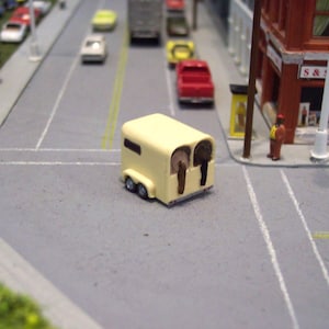 Puede incluir: Un modelo en miniatura de un remolque para caballos, pintado de color amarillo claro, está situado en una calle gris. El remolque tiene dos cabezas de caballo visibles. La escena incluye coches en miniatura, un edificio con el letrero "S & Drugs" y una figura.
