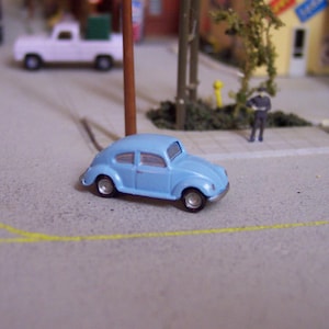 May include: A light blue toy car, a miniature Volkswagen Beetle, is positioned on a gray surface, possibly a model of a street. The car has silver wheels and is in front of a miniature town scene.