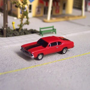 Op de afbeelding: Een miniatuur rode klassieke auto met zwarte racestrepen. De speelgoedauto staat op een grijze ondergrond, met een gele lijn en een miniatuur straatbeeld op de achtergrond. De auto heeft zilveren wielen.