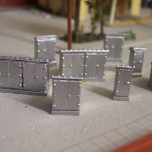 May include: Miniature silver metal electrical boxes, likely for a model train or diorama. The boxes have a rectangular shape with two doors and are decorated with small rivets. A small figure in a red hat and brown coat stands nearby.