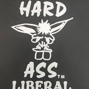 May include: Black background with white text that reads "HARD ASS LIBERAL" with a cartoon character in the center. The character has large ears and a spiky head.