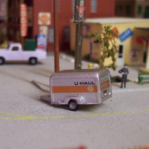 Puede incluir: Un pequeño remolque U-Haul plateado con rayas naranjas y el logotipo de U-Haul está estacionado sobre una superficie gris. El remolque tiene una rueda visible y está enganchado a un enganche. La escena incluye edificios en miniatura y una camioneta blanca.