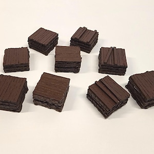 May include: Nine small, square, wooden blocks with a dark brown finish are arranged on a white surface. The blocks have a layered appearance, with some showing individual planks.