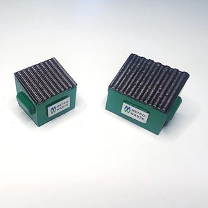 Peut inclure: Deux mini-conteneurs verts avec des toits noirs ondulés. Chacun a une petite poignée et une étiquette blanche avec le texte "METRO WASTE" et un logo. Les conteneurs sont posés sur un fond blanc.