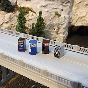 May include: Miniature vending machines in various colors, including blue, black, and silver, are arranged on a model bridge. The machines have detailed graphics and are set against a backdrop of a model landscape with trees and a tunnel.