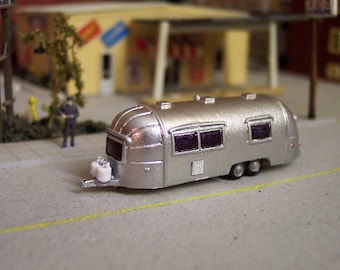 Może przedstawiać: Srebrna miniaturowa przyczepa kempingowa o zaokrąglonym, aerodynamicznym kształcie, z ciemnofioletowymi oknami, podwójnymi kołami i dwiema małymi białymi butlami z propanem zamontowanymi na przednim zaczepie, jako szczegółowy model kolekcjonerski.
