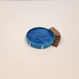 May include: A miniature swimming pool with a blue interior and a brown deck. The pool is round with a light blue exterior and white supports. The water inside the pool is a darker blue, with a rippled texture.