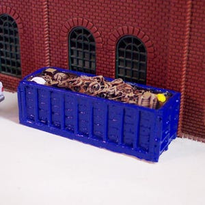 Peut inclure: Une miniature benne bleue remplie de débris est placée devant un bâtiment en briques avec des fenêtres en arc. Une petite camionnette blanche avec un objet vert dans la benne est garée à proximité. La scène est une maquette ou un diorama.
