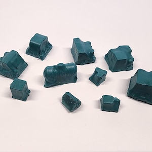May include: A collection of teal-colored, irregularly shaped wax melts. The wax pieces vary in size and shape, resembling small, textured rocks. They are arranged on a white surface.