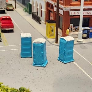 May include: Miniature blue portable toilets with white roofs are positioned on a gray street. Toy cars and buildings are in the background. The scene is a diorama or model of a city street.