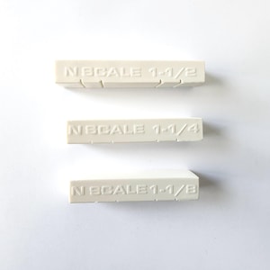 May include: Three off-white rectangular model train parts are arranged on a white surface. Each part has the text "N SCALE" followed by a fraction: 1-1/2, 1-1/4, and 1-1/8. The parts have grooves on the sides.