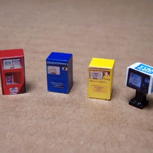 Puede incluir: Miniaturas de quioscos de periódicos en rojo, azul y amarillo, con un pequeño televisor que muestra "USA TODAY". Los quioscos tienen gráficos y texto detallados, lo que sugiere un modelo o diorama. Los objetos están sobre un fondo neutro.