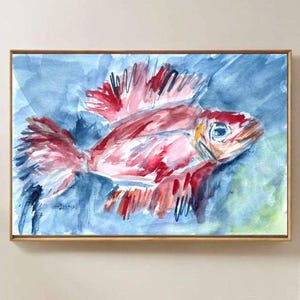 Puede incluir: Pintura a la acuarela de un pez rojo y rosa con una aleta grande, sobre un fondo azul. La obra de arte está enmarcada en un marco dorado. El pez tiene un ojo detallado y es el punto focal de la pintura.