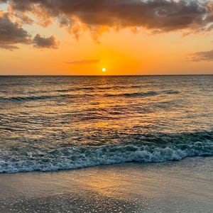 May include: A scenic view of a sunset over the ocean. The sun is a bright yellow orb, casting a golden glow on the water. The sky is a mix of orange, yellow, and gray clouds. Gentle waves roll onto the sandy beach.