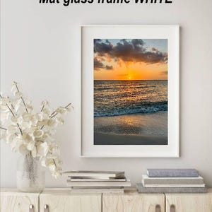 May include: White mat glass frame with a sunset beach scene. The image features a vibrant orange and yellow sunset over the ocean, with waves gently lapping the shore. The text "Mat glass frame WHITE" is at the top.