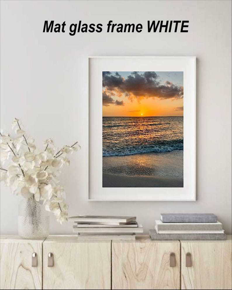 May include: White mat glass frame with a sunset beach scene. The image features a vibrant orange and yellow sunset over the ocean, with waves gently lapping the shore. The text "Mat glass frame WHITE" is at the top.