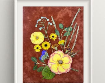 May include: A framed floral artwork featuring a vibrant arrangement of pressed flowers. The composition includes yellow roses, sunflowers, and blue blossoms against a textured, reddish-brown background. The white frame enhances the artwork's presentation.