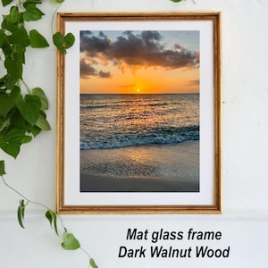 May include: A framed print of a sunset over the ocean. The image features a golden frame, a white mat, and a beach scene with a vibrant orange and yellow sky. The text "Mat glass frame Dark Walnut Wood" is visible.
