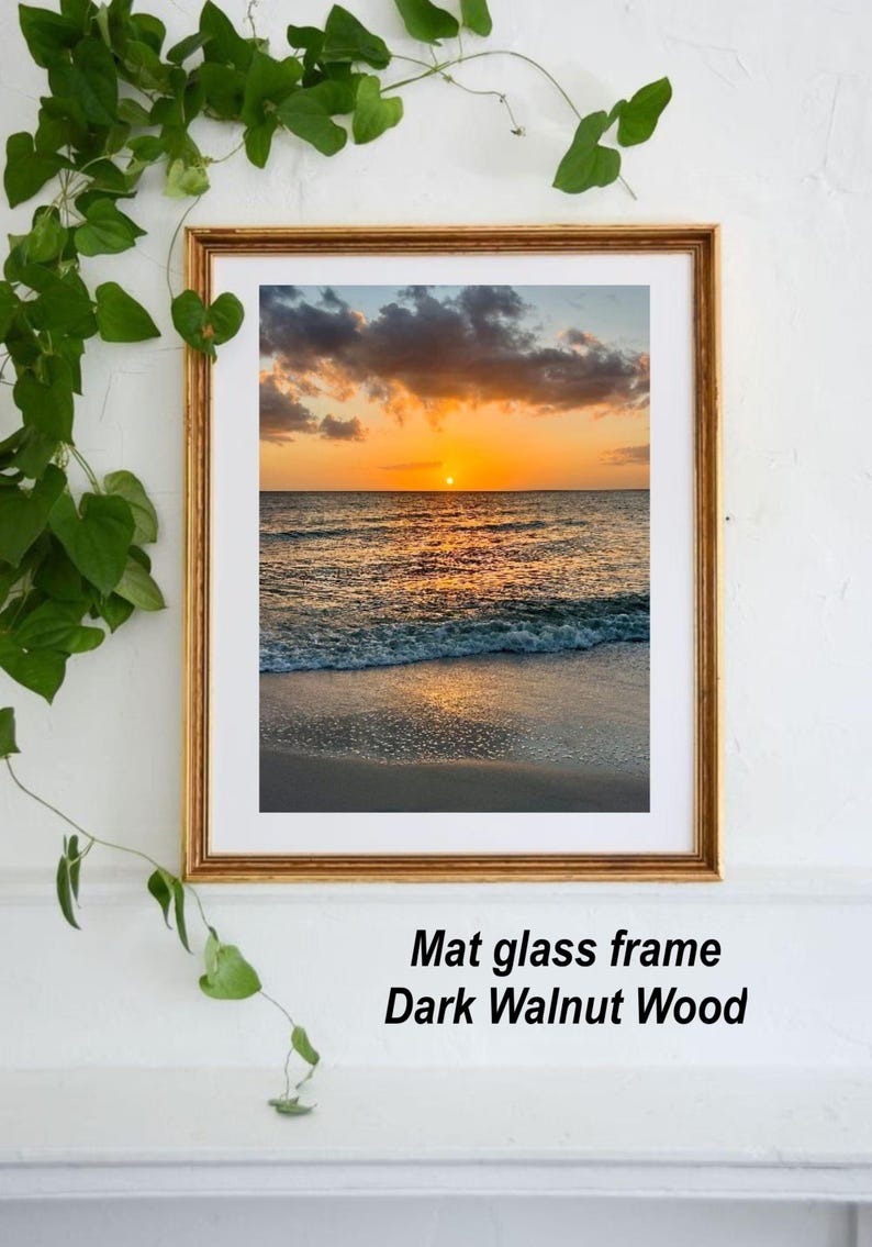 May include: A framed print of a sunset over the ocean. The image features a golden frame, a white mat, and a beach scene with a vibrant orange and yellow sky. The text "Mat glass frame Dark Walnut Wood" is visible.