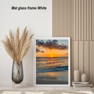 May include: A framed print of a sunset over the ocean, with a white mat and frame. The image shows a vibrant orange sun setting over the water, with waves lapping the shore. The text "Mat glass frame White" is visible.