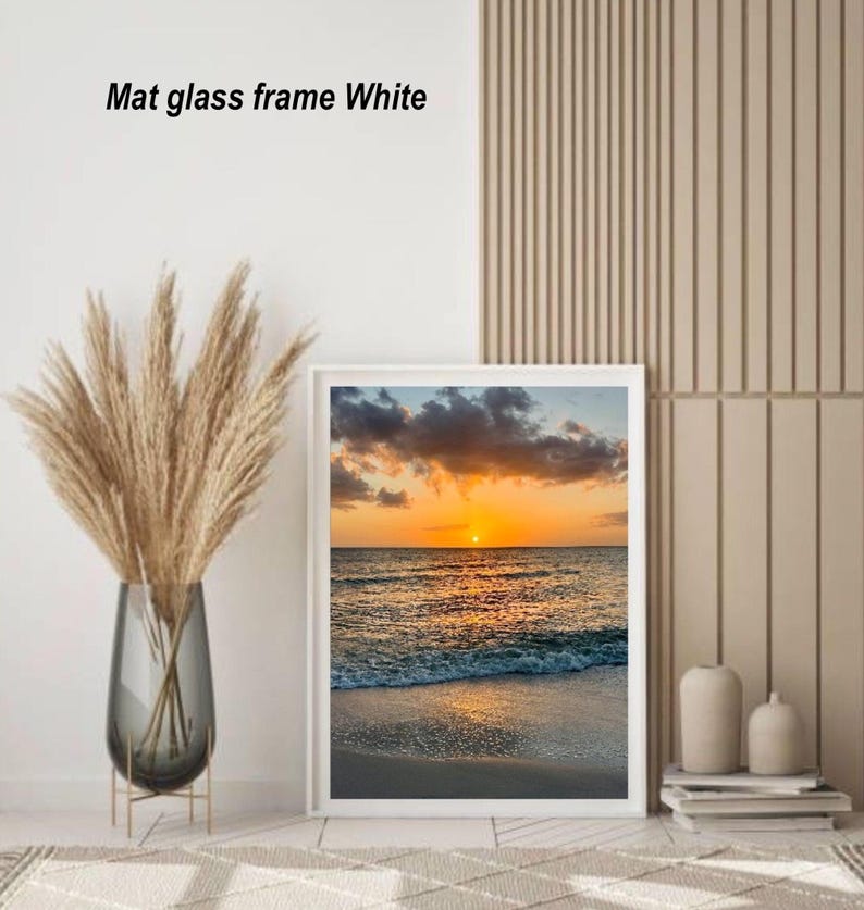 May include: A framed print of a sunset over the ocean, with a white mat and frame. The image shows a vibrant orange sun setting over the water, with waves lapping the shore. The text "Mat glass frame White" is visible.
