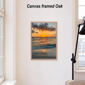 May include: A framed canvas print of a sunset over the ocean. The image features a vibrant orange and yellow sky, with clouds and the sun reflecting on the water. The frame is made of light-colored wood, and the text "Canvas framed Oak" is above the image.