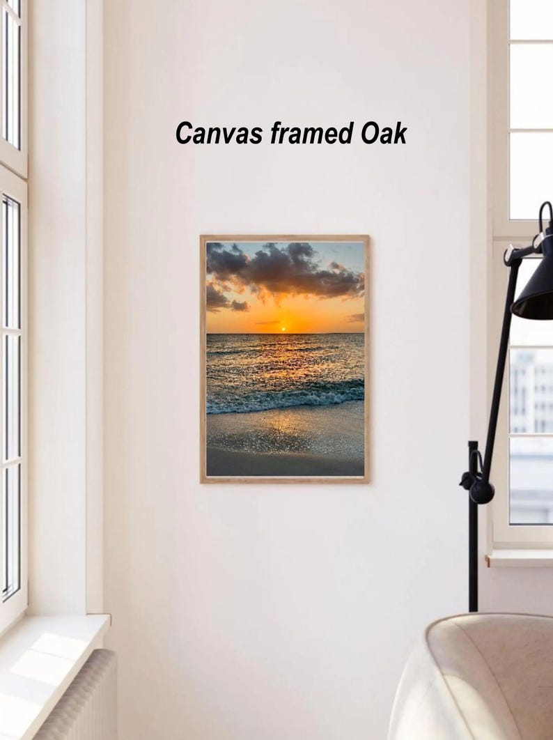 May include: A framed canvas print of a sunset over the ocean. The image features a vibrant orange and yellow sky, with clouds and the sun reflecting on the water. The frame is made of light-colored wood, and the text "Canvas framed Oak" is above the image.
