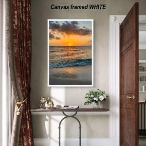 May include: A framed canvas print with a sunset over the ocean. The image is in a white frame and the text "Canvas framed WHITE" is above the frame. The scene includes a beach, water, and a vibrant orange and yellow sky.