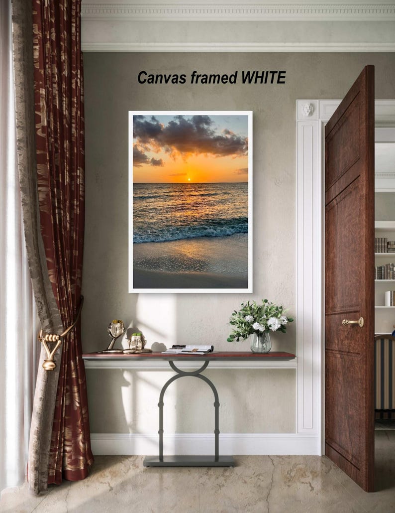 May include: A framed canvas print with a sunset over the ocean. The image is in a white frame and the text "Canvas framed WHITE" is above the frame. The scene includes a beach, water, and a vibrant orange and yellow sky.
