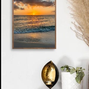 May include: A framed canvas print of a sunset over the ocean. The sky is filled with orange and yellow hues, and the water reflects the sunlight. The frame is a dark wood. Text on the top reads "Canvas framed Maple".