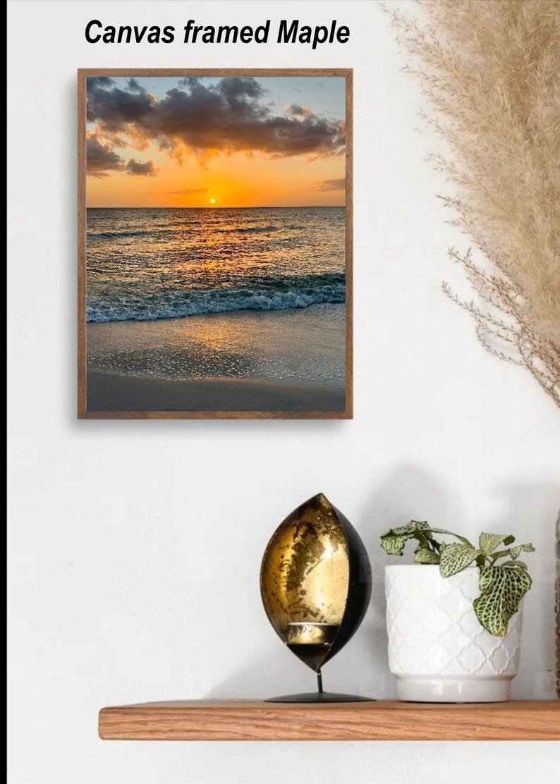 May include: A framed canvas print of a sunset over the ocean. The sky is filled with orange and yellow hues, and the water reflects the sunlight. The frame is a dark wood. Text on the top reads "Canvas framed Maple".