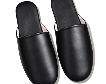 May include: A pair of black slip-on slippers featuring a smooth, solid-colored exterior and a light-colored interior lining. The footwear has a rounded toe design and an open-back construction, presented from a top-down perspective.