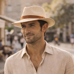 Puede incluir: Un hombre con un sombrero fedora beige con una banda marrón, una camisa beige y pantalones color caqui. Sostiene una taza de café blanca y camina por una calle. El texto dice: "Not just stylish. He'll use it. Always."